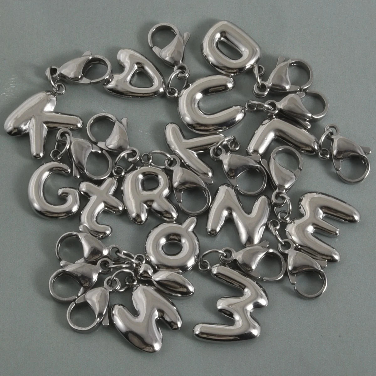 A collection of stainless steel alphabet letter charms with lobster clasps arranged on a gray surface.