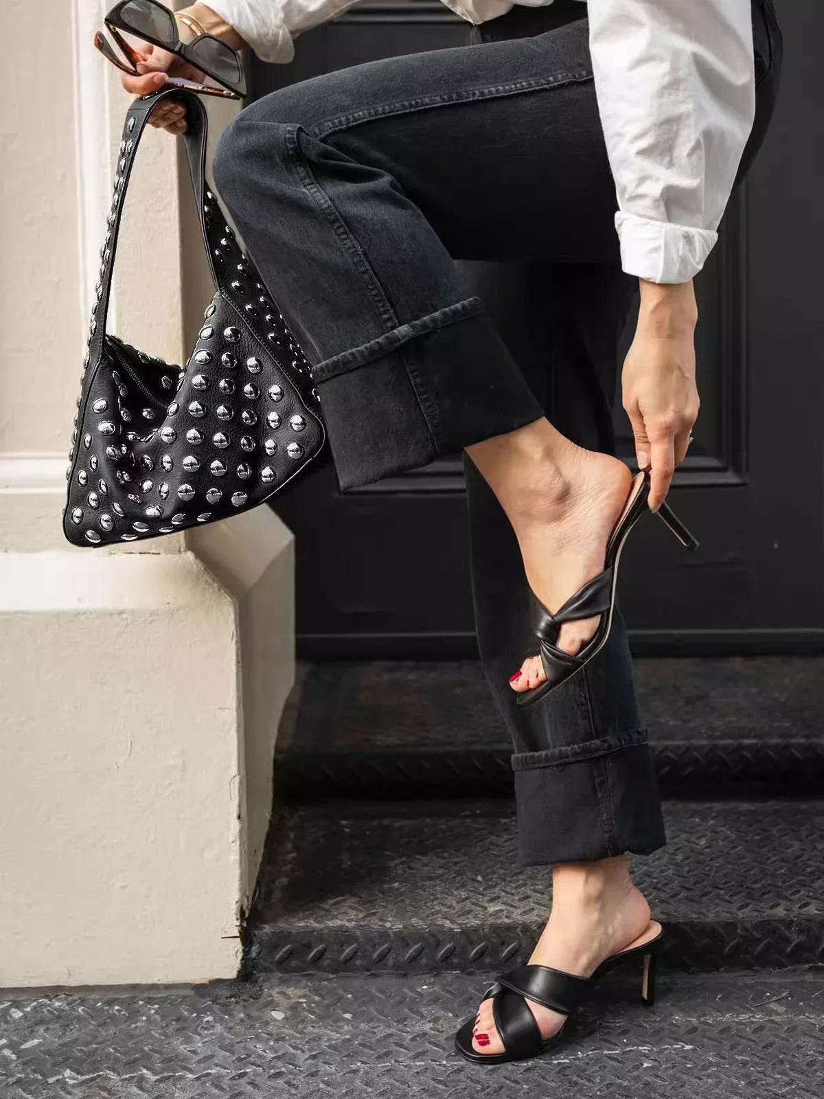 A person wearing dark pants is holding a black studded shoulder bag while standing on steps.