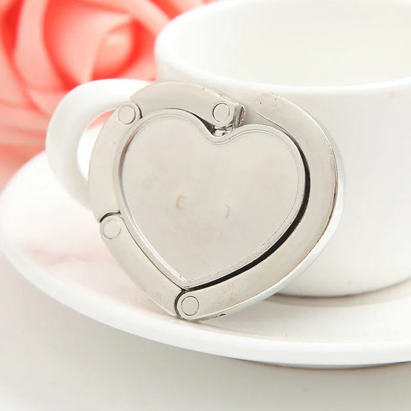 A silver heart-shaped folding alloy handbag hanger hook resting on a white saucer next to a white cup.