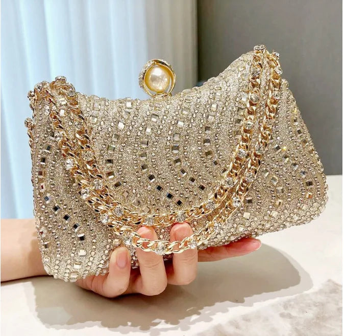 A hand holds a sparkling gold and diamond-inlay evening clutch bag with a pearl closure.