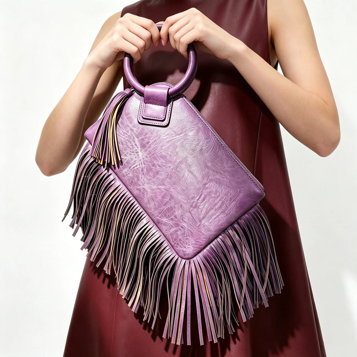 A hand holds a purple leather fringed clutch bag with tassels against a dark maroon dress.