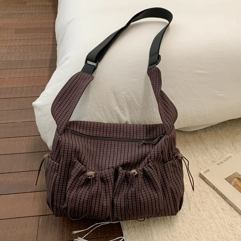 A dark brown plaid nylon drawstring tote bag resting on a white pillow next to a wooden floor.