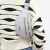 A woman wearing a cream striped sweater and blue jeans is holding a light gray nylon crossbody fanny pack.