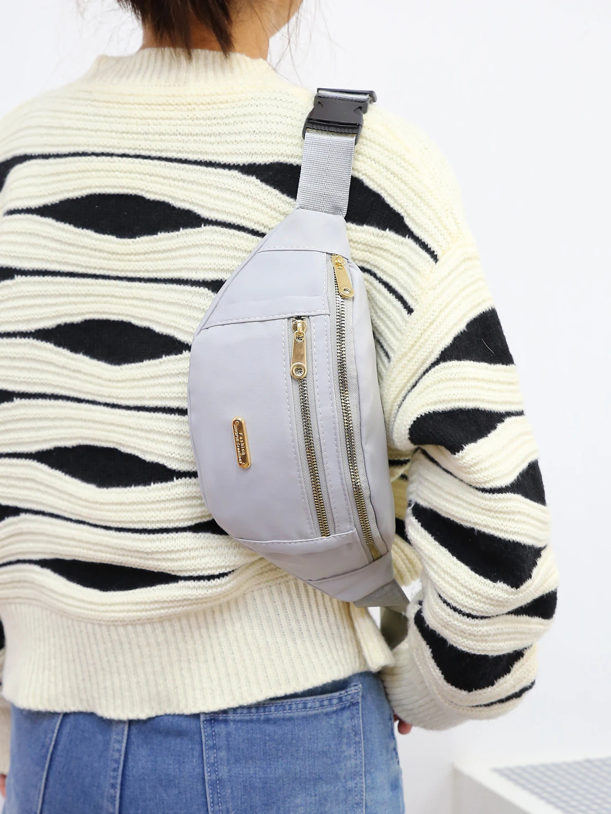 A woman wearing a cream striped sweater and blue jeans is holding a light gray nylon crossbody fanny pack.