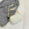 A beige nylon cross-body waist bag with a light tan strap is laid on a white surface next to a gray denim jacket.