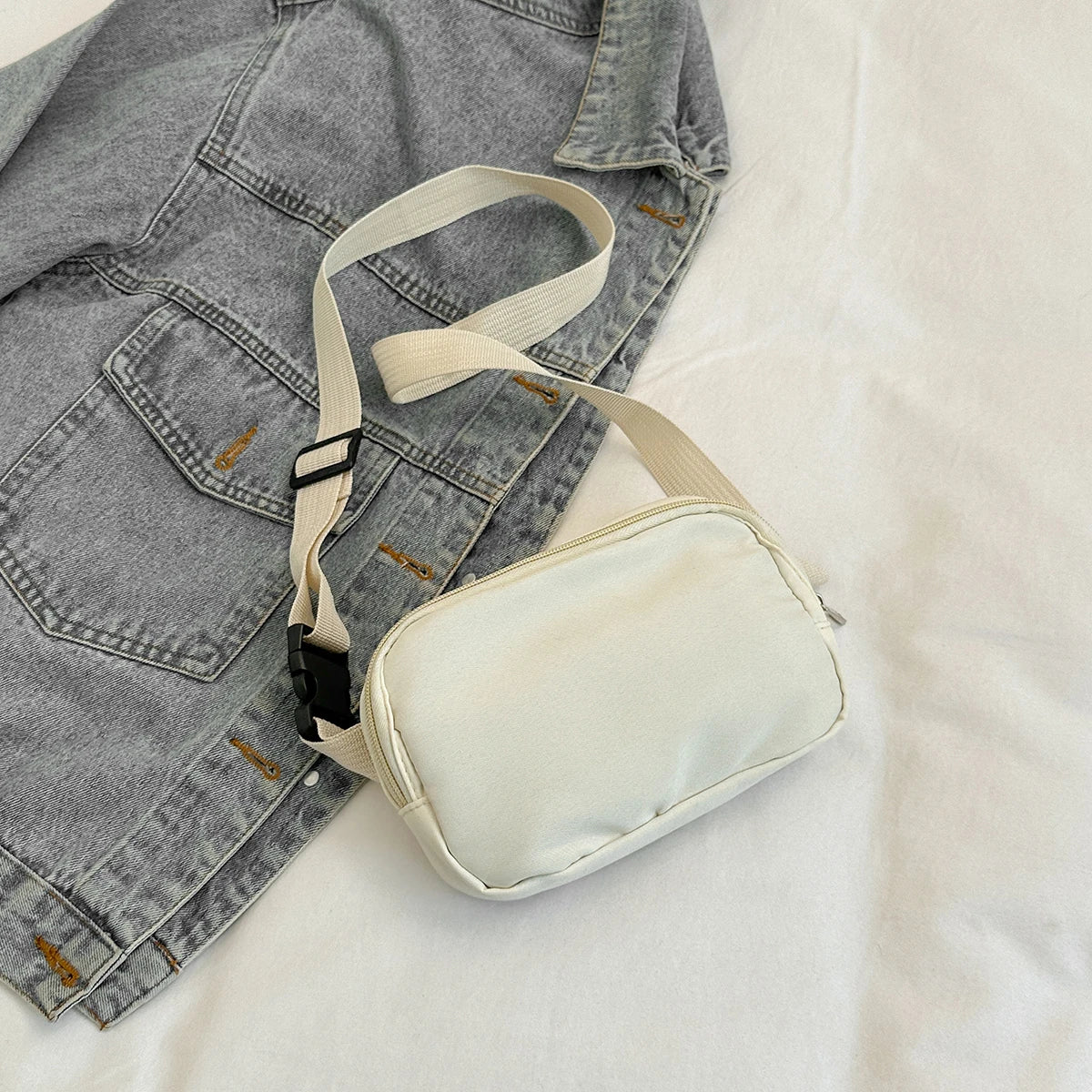 A beige nylon cross-body waist bag with a light tan strap is laid on a white surface next to a gray denim jacket.