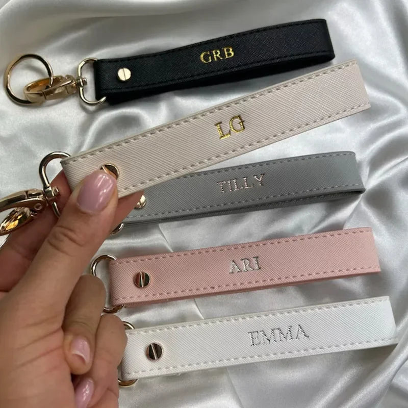 A hand holds several personalized leather wristlet keychains with gold hardware and various names engraved on them.