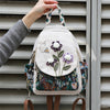 A handcrafted linen backpack featuring light beige fabric and purple floral embroidery, held by a hand against a gray slatted