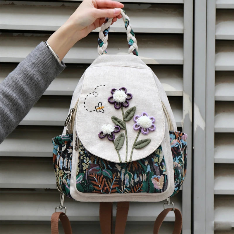 A handcrafted linen backpack featuring light beige fabric and purple floral embroidery, held by a hand against a gray slatted