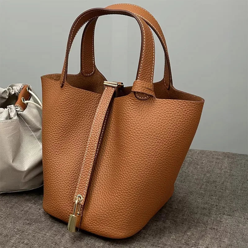 A tan genuine leather bucket bag with a flap closure and gold hardware resting on a gray surface.