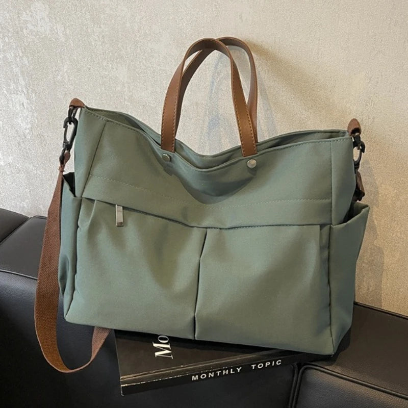 A light sage green nylon tote bag with brown straps and multiple internal pockets resting on a dark surface.