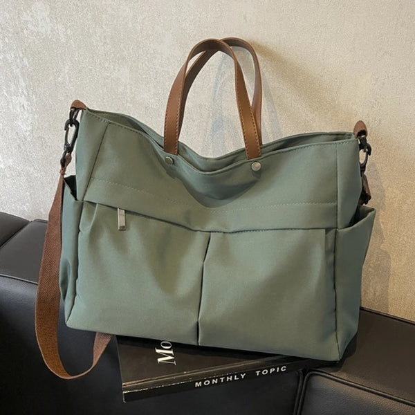 A light sage green nylon tote bag with brown straps and multiple internal pockets resting on a dark surface.