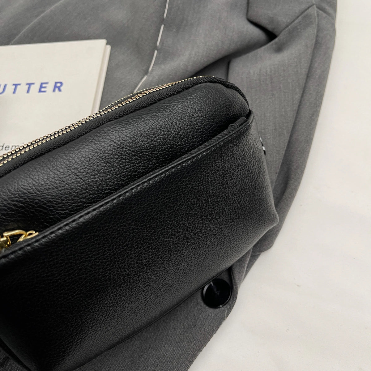 A close-up of a black vegan PU crossbody bag with a gold zipper on a gray fabric background.