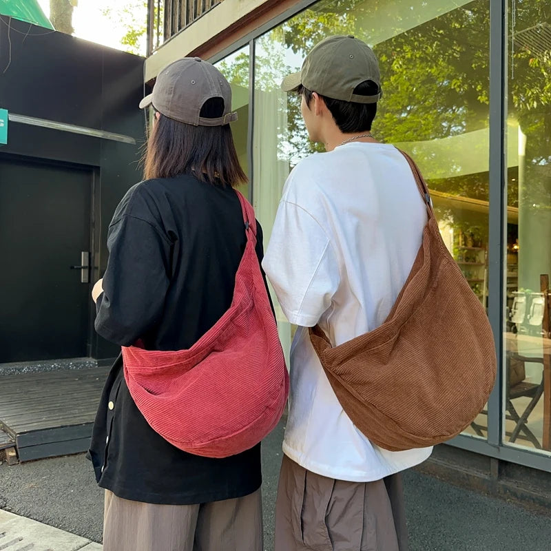 A person wearing a black shirt and a grey cap holds a reddish-pink corduroy casual crossbody bag in front of a glass door.