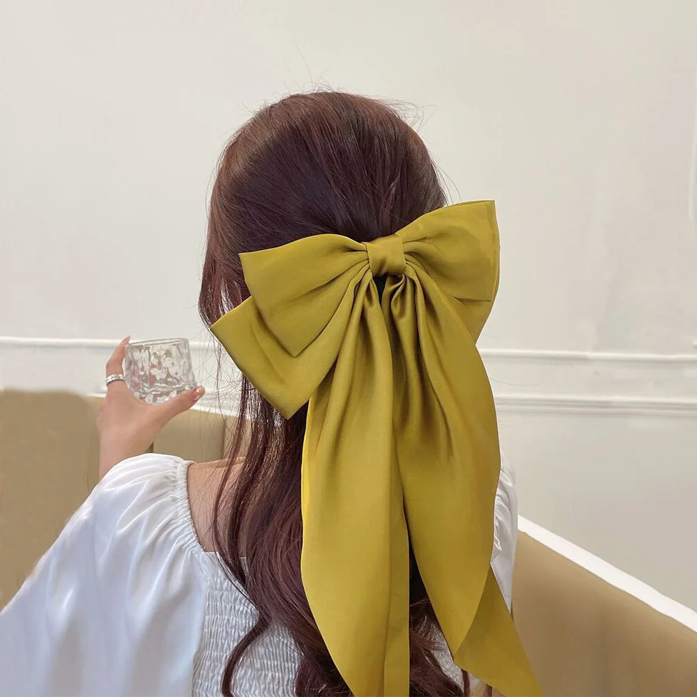 A woman with long brown hair is shown from the back, wearing a large olive green silk bow tied in her hair.