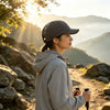 A woman wearing a gray hoodie and black mesh baseball cap stands outdoors with mountains in the background during sunset.