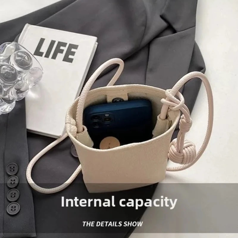 Bolso cruz de cuero beige con un smartphone azul, apoyado sobre una tela gris y un libro.