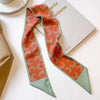 Un foulard pour cheveux en soie floral élégant aux motifs orange et vert, à côté d'un carnet blanc.