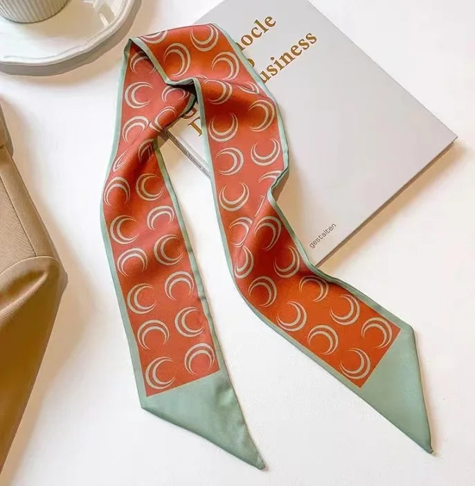 Un foulard pour cheveux en soie floral élégant aux motifs orange et vert, à côté d'un carnet blanc.