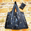 A black reusable shopping bag with gold leaf accents is displayed against a textured brick background.