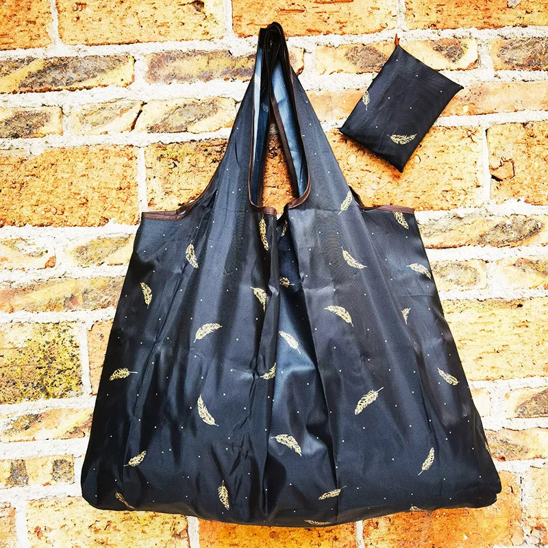 A black reusable shopping bag with gold leaf accents is displayed against a textured brick background.