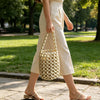 A woman wearing a cream-colored dress and light trousers walks outdoors carrying an elegant beaded clutch bag.