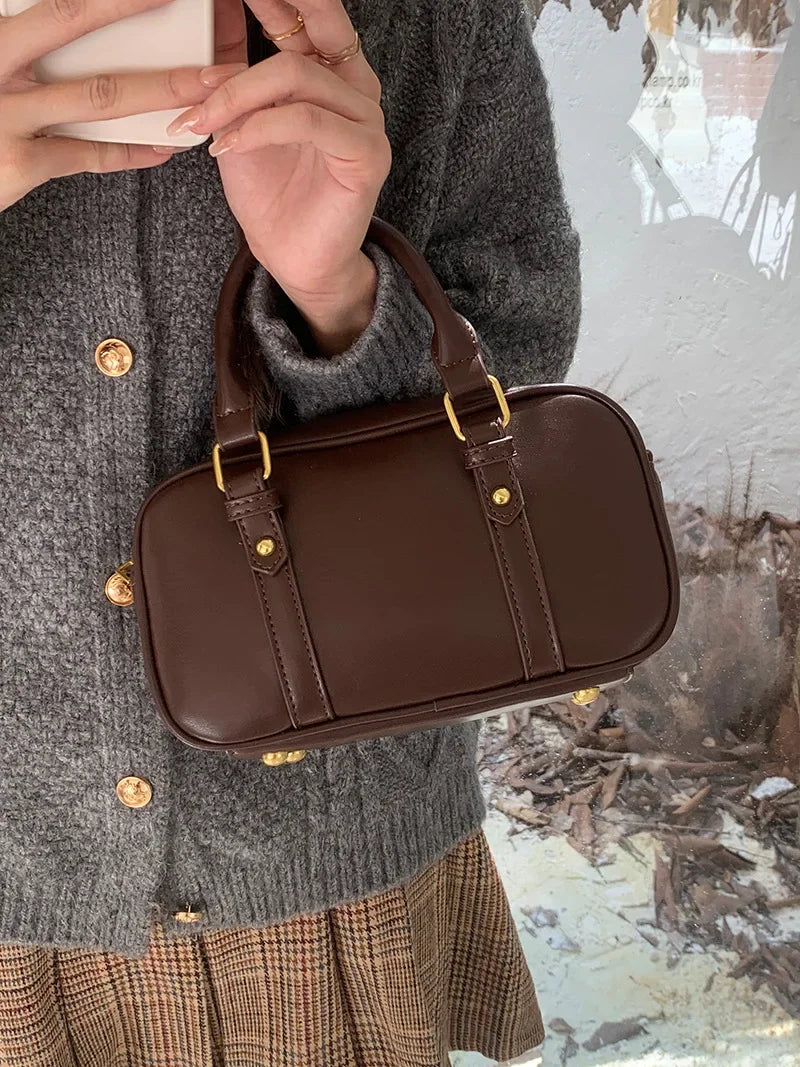 A brown vintage PU leather shoulder bag is held by a person wearing a gray sweater and plaid skirt.