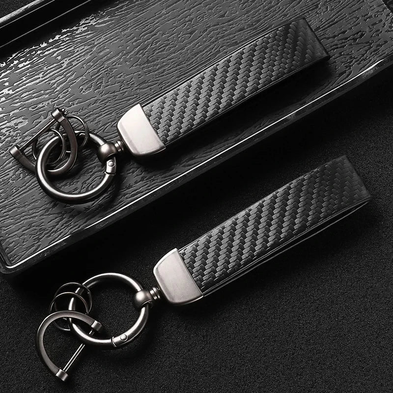 A close-up shot of a black carbon fiber leather keychain with silver hardware resting on a dark surface.