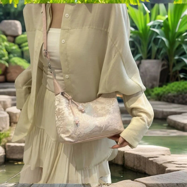 A woman wearing a light beige dress holds a cream embroidered crossbody shoulder bag in an outdoor setting.