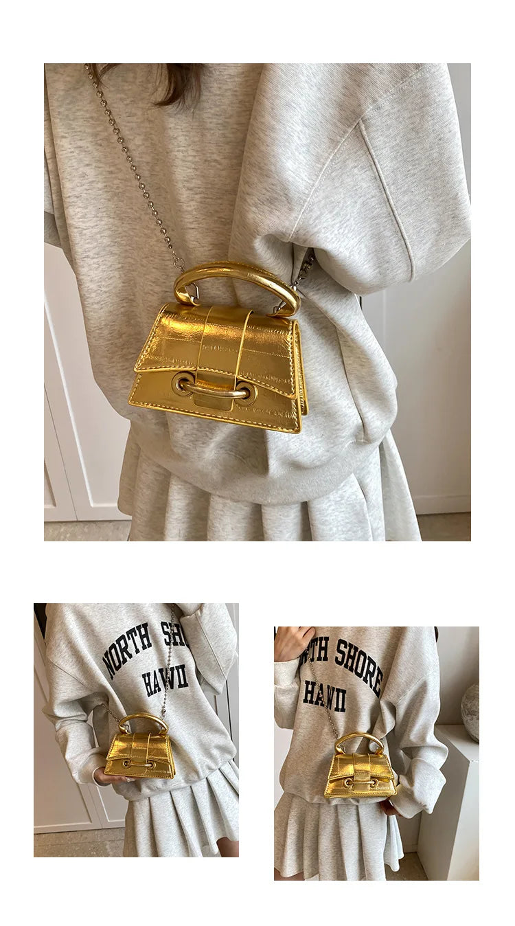A gold square crossbody bag with a chain strap is shown hanging from a sleeve of a light gray sweater.
