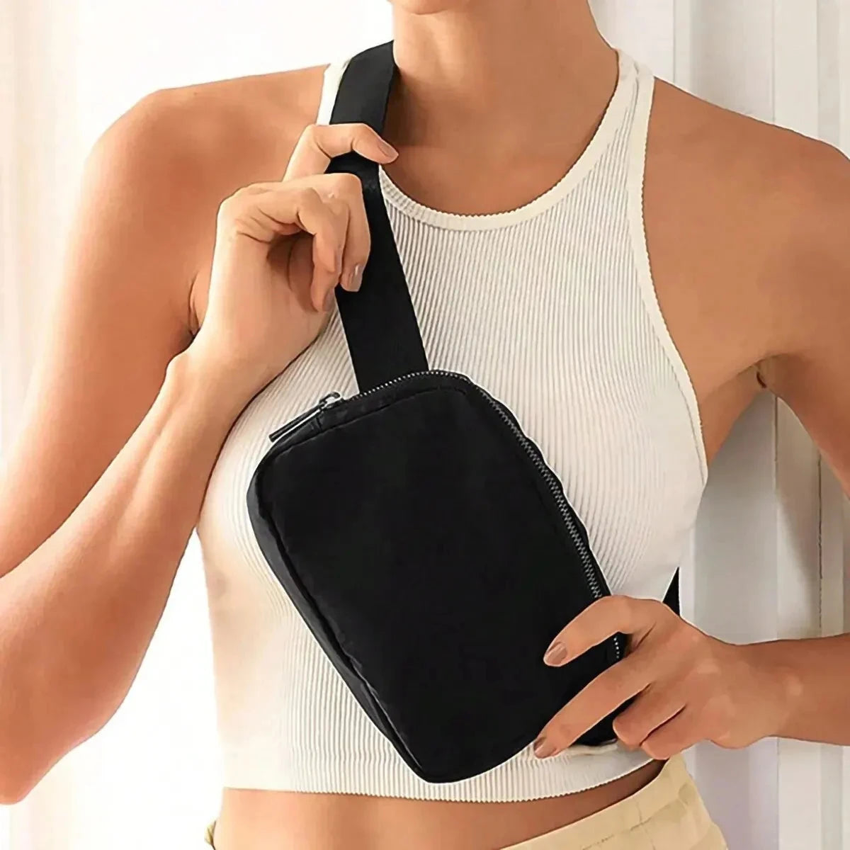 A woman wearing a white ribbed tank top is holding a black cross-body nylon waist bag.