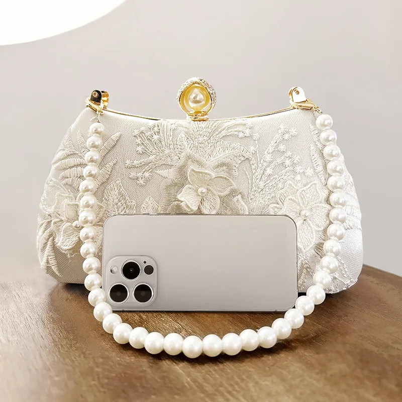 A white embroidered pearl chain clutch bag with a clear phone case resting on it.