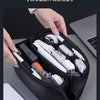 A hand is opening a black toiletry bag organizer revealing various electronic cables and accessories.