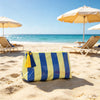 A yellow and blue striped cosmetic travel pouch with yellow fringe lying on a sandy beach with beach chairs in the background