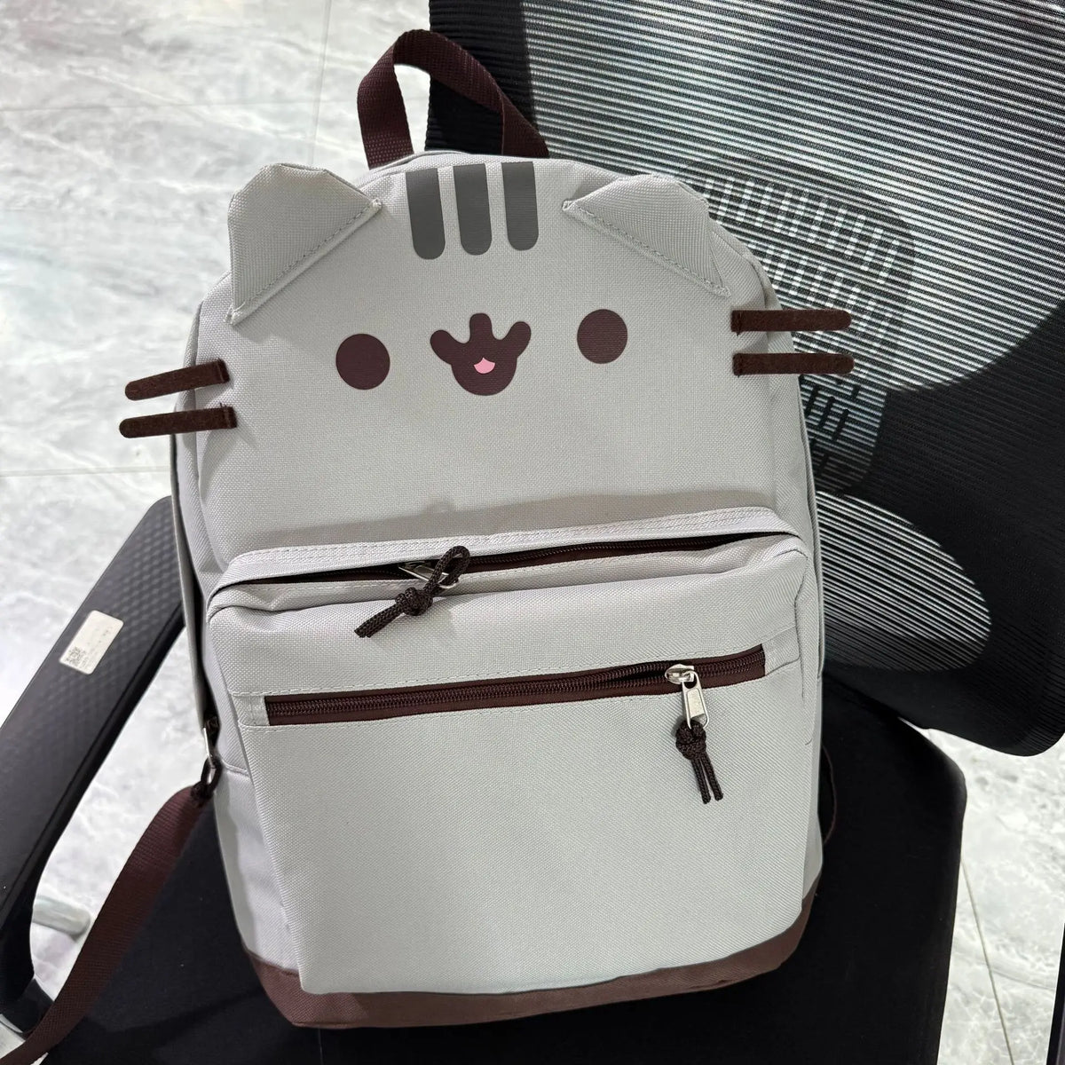 A light gray Pusheen cat canvas backpack with brown accents resting on a black chair.