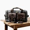 Dunkelgrabe Reise-Gepäcktasche aus Canvas mit braunen Lederakzenten auf Holzoberfläche.