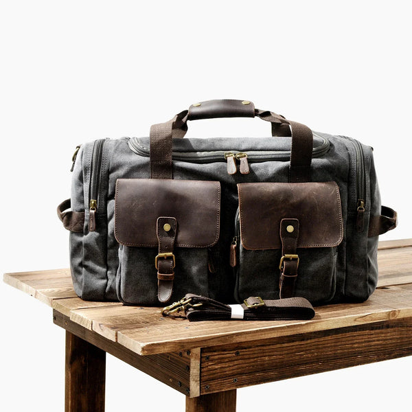 Dunkelgrabe Reise-Gepäcktasche aus Canvas mit braunen Lederakzenten auf Holzoberfläche.