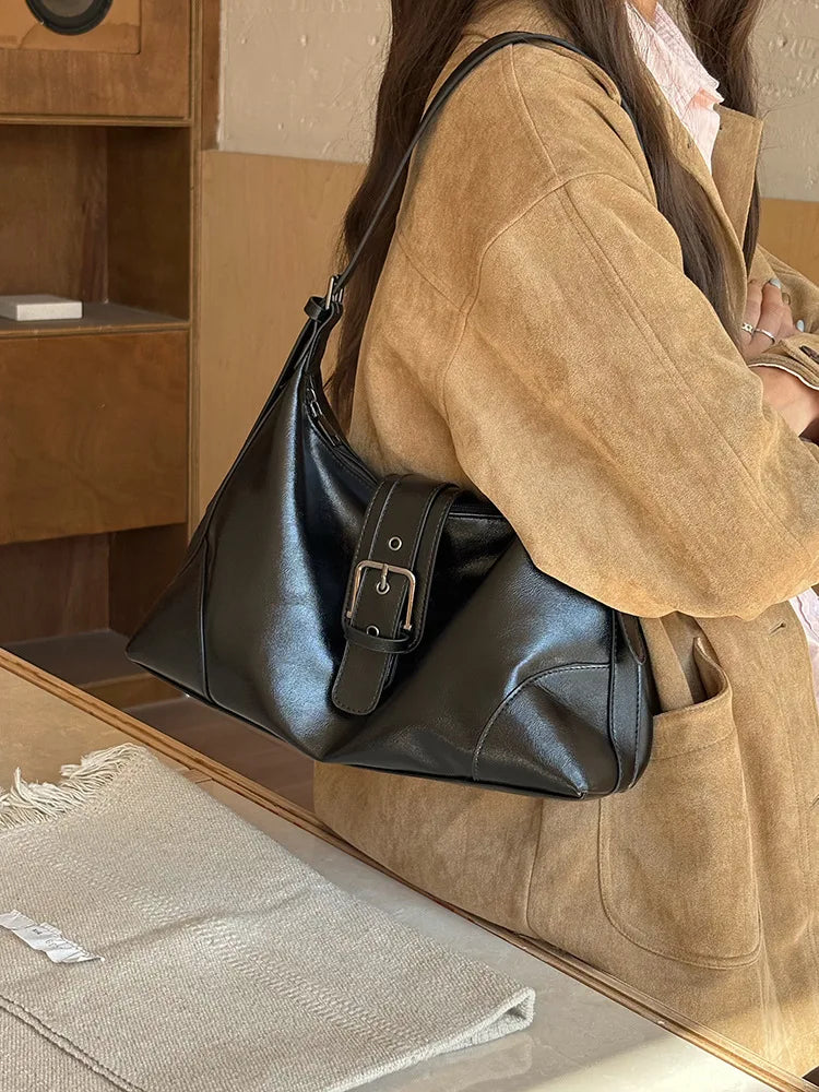 A woman holds a black PU leather underarm bag in a tan jacket, resting on a glass surface.