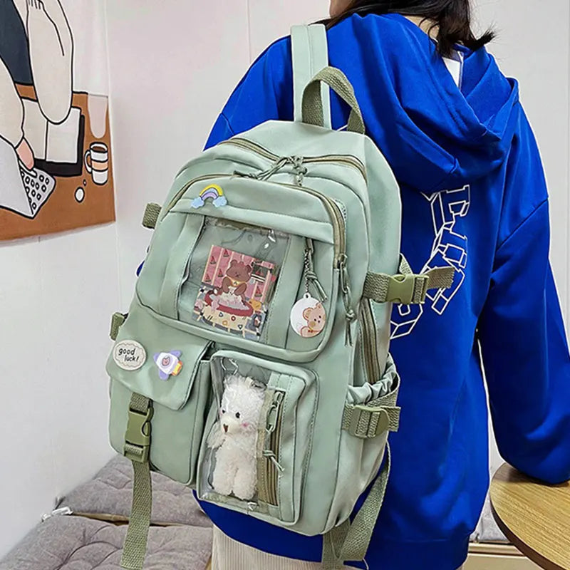 A woman is wearing a light green backpack with clear pockets and a fluffy white charm against a blue hoodie.