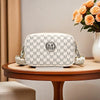 A white crossbody bag with a gray M-pattern print and silver hardware resting on a wooden table.