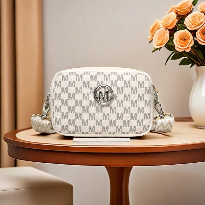A white crossbody bag with a gray M-pattern print and silver hardware resting on a wooden table.