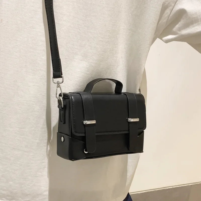 A black minimalist leather crossbody bag is shown hanging on a white shirt.