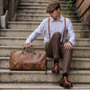 Ein Mann in blauem Hemd und braunen Hosen sitzt auf Steinstufen und hält eine vintage geprägte Leder-Tasche.