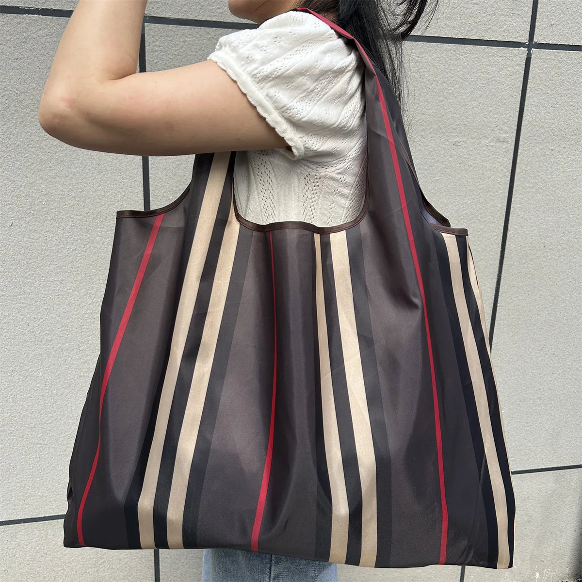 A person is holding a brown and beige striped foldable grocery tote bag against a light gray wall.