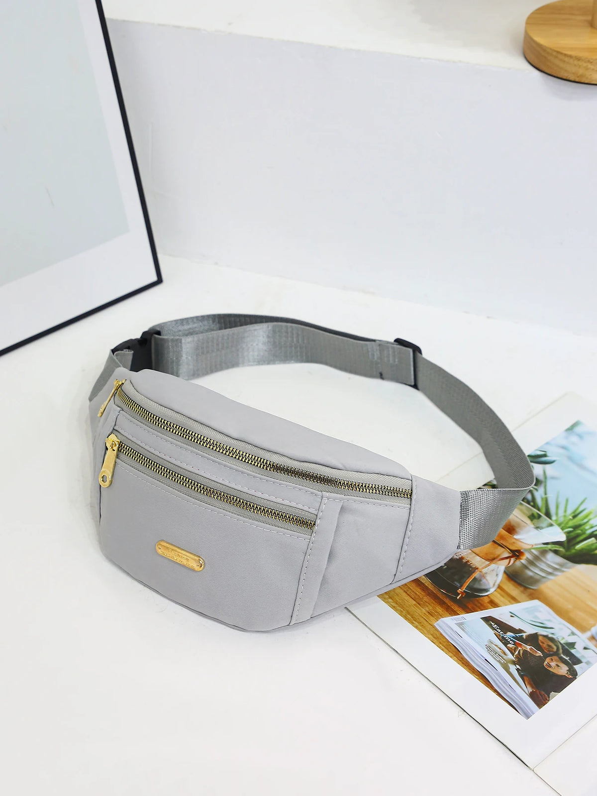 A light gray nylon crossbody fanny pack with a gold zipper and leather accent resting on a white surface.