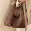A brown suede shoulder tote bag carried by a person wearing a beige jacket and white pants.