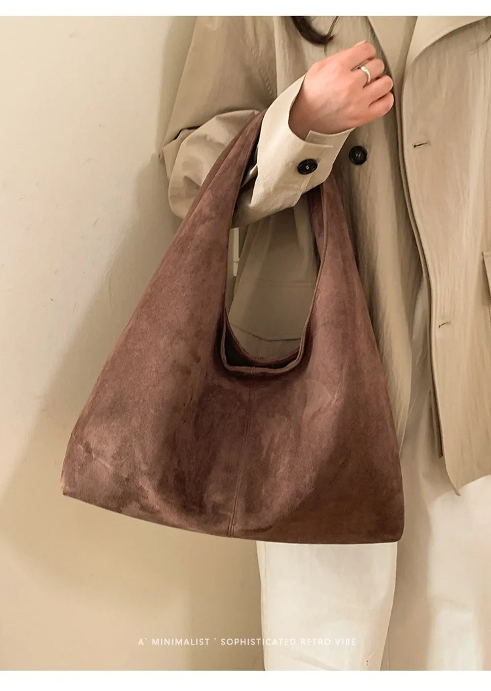 A brown suede shoulder tote bag carried by a person wearing a beige jacket and white pants.