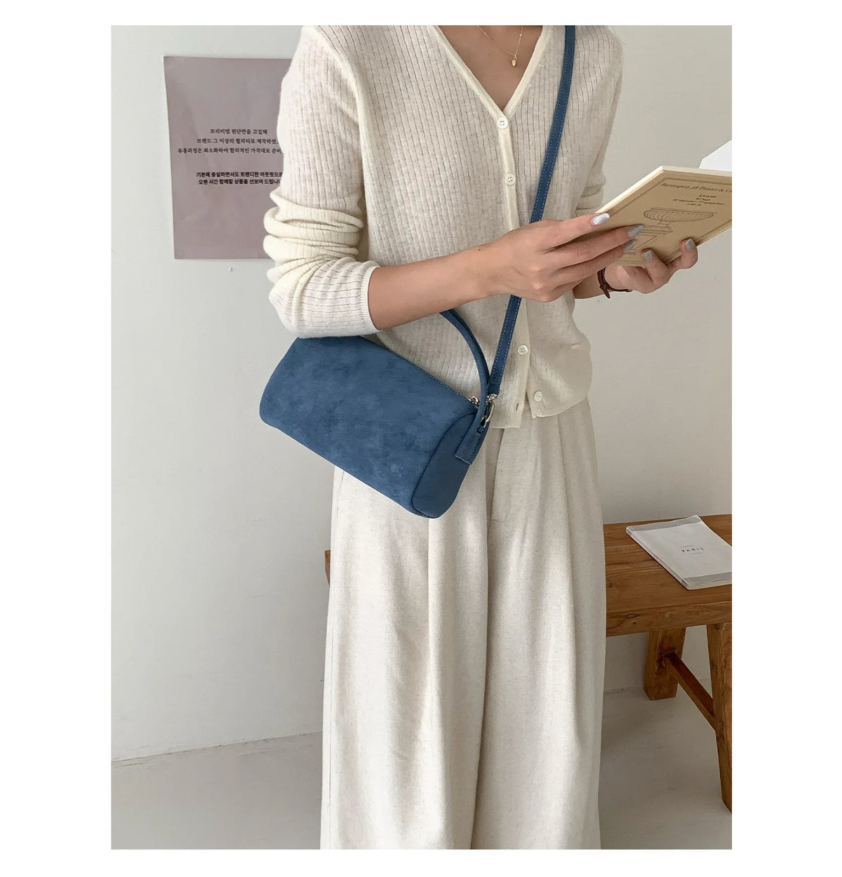 A woman wearing a cream knit dress holds a navy blue faux suede crossbody bag and a book.