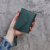 A hand holds a dark green PU leather coin clutch wallet with a gold tassel against a gray background.