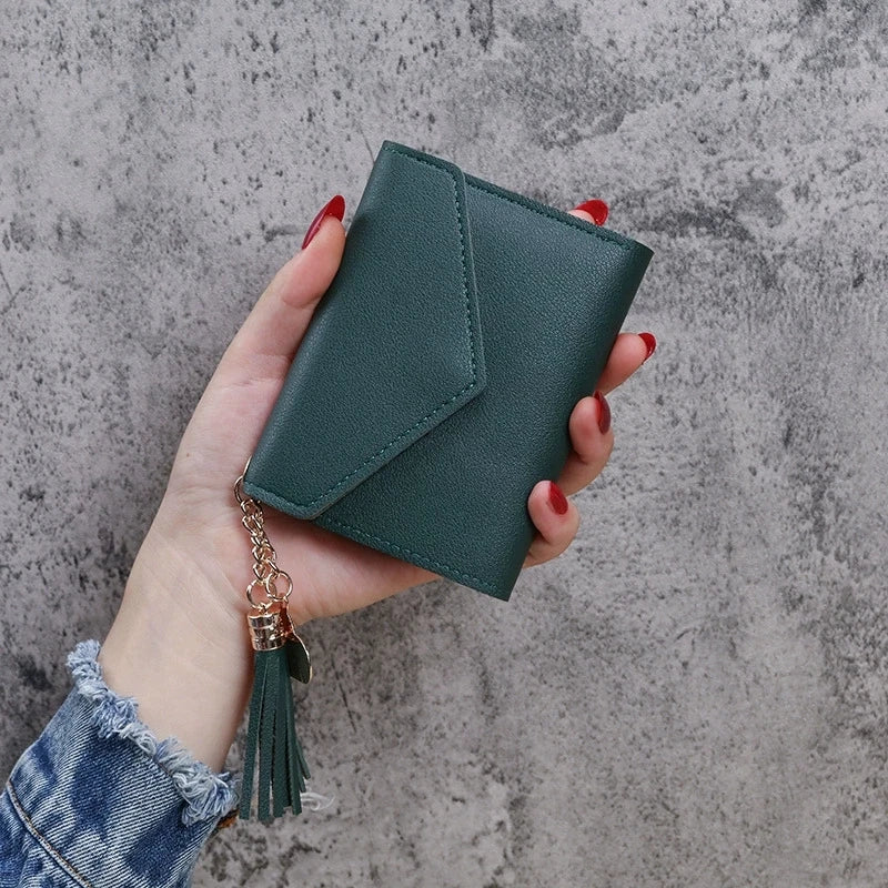 A hand holds a dark green PU leather coin clutch wallet with a gold tassel against a gray background.